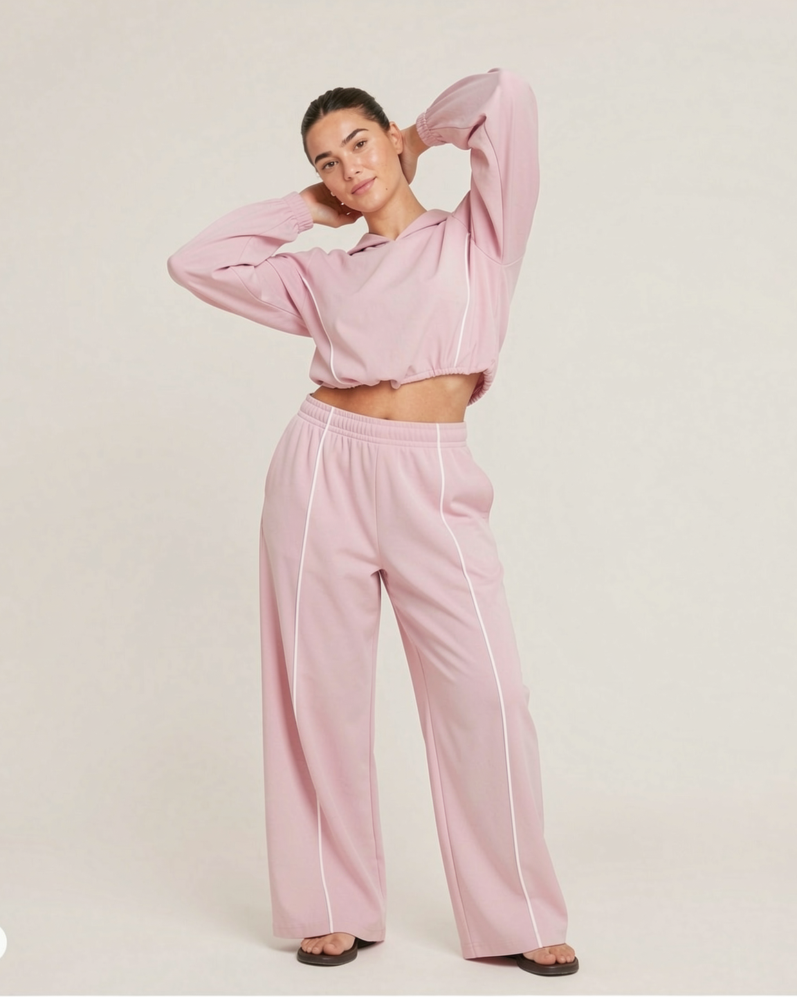 A woman modeling a light pink two-piece lounge set consisting of a cropped long-sleeve top and matching wide-leg pants with thin white side stripes, set against a plain light beige studio background.