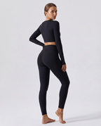 A woman modeling a matching black long-sleeve crop top and high-waisted leggings set, shown from a three-quarters back angle against a light gray studio background.